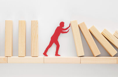 Houten blokjes op een rij, deel dreigt om te vallen maar worden tegengehouden door een figuur