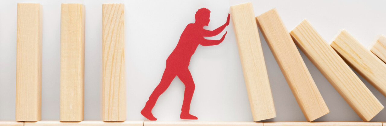 Houten blokjes op een rij, deel dreigt om te vallen maar worden tegengehouden door een figuur
